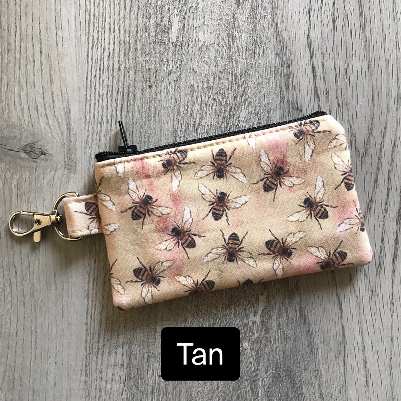 May include: Tan zippered pouch with a bee print. The pouch has a black zipper and a silver key ring.