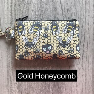 May include: A small zippered pouch with a gold honeycomb pattern and black bees. The pouch has a black zipper and a black ring for attaching a key chain. The text "Gold Honeycomb" is printed below the pouch.