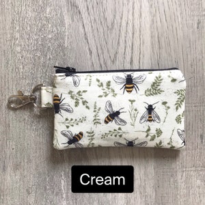 May include: A cream-colored zippered pouch with a black zipper and a black metal key ring. The pouch has a repeating pattern of bees and green foliage.