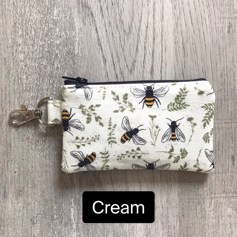 May include: A cream-colored zippered pouch with a black zipper and a black metal key ring. The pouch has a repeating pattern of bees and green foliage.