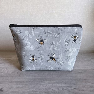 May include: A gray zippered pouch with a repeating pattern of bees and floral sprigs. The pouch is made of a canvas-like material.