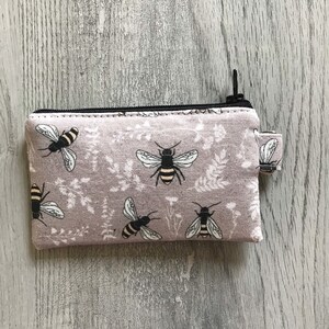 May include: A small zippered pouch with a light purple background and a repeating pattern of black and yellow bees. The pouch has a black zipper and a black pull tab.