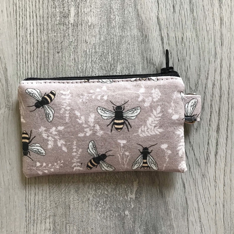 May include: A small zippered pouch with a light purple background and a repeating pattern of black and yellow bees. The pouch has a black zipper and a black pull tab.