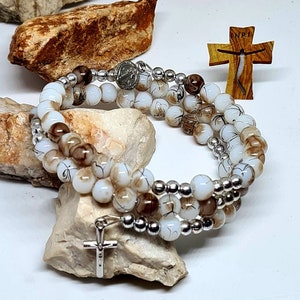 Puede incluir: Un brazalete de triple hebra con cuentas de vidrio blancas y marrones, separadores plateados y un dije de cruz plateada. El brazalete está sobre una superficie blanca con piedras.