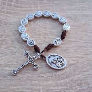 May include: A silver-toned rosary with oval beads featuring religious figures, connected by brown cord. A detailed cross and a pendant with the text "CARMEL VIRGIN ITALY" are also present. The rosary is displayed on a light wooden surface.