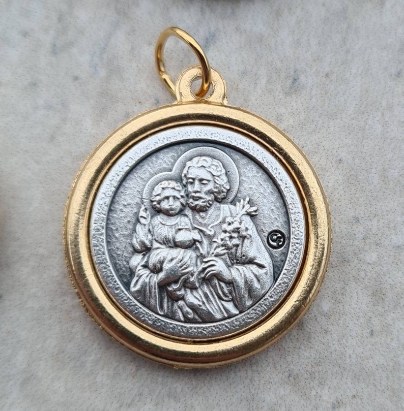 May include: A gold-toned pendant featuring a silver-toned image of Saint Anthony of Padua holding the Christ Child. The pendant has a loop at the top for attaching to a chain.