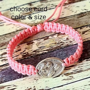 May include: A pink braided cord bracelet with a silver charm featuring a depiction of Saint Ann. The charm has the words "St. Ann" inscribed on it. The bracelet is on a wooden surface. Choose cord colour and size.