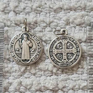 Puede incluir: Dos medallas de San Cristóbal de plata. Una medalla presenta una representación de San Cristóbal llevando a un niño, con la inscripción "SNTIA MUNIAMVR" alrededor del borde. La otra medalla presenta una cruz con la inscripción "PAX VRSN" alrededor del borde.