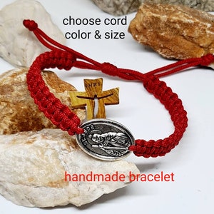 Könnte beinhalten: Rotes geflochtenes Armband mit einem silbernen Anhänger, der eine religiöse Figur und die Worte "St. Jude" zeigt. Das Armband enthält auch ein Holzkreuz mit der Inschrift "INRI".
