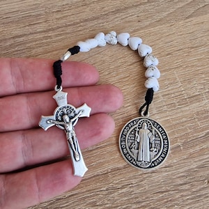 May include: A silver rosary with a black cord, white heart-shaped beads, a silver crucifix with the inscription "INRI", and a silver medal with the image of Saint Anthony of Padua. The medal has the inscription "TV-NRO-PRA EIVS-IN-OB/ SENTIA MUNIAM MVR".