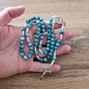 Könnte beinhalten: Ein blaues und weißes Rosenkranz mit einem silbernen Kreuz und einem silbernen Anhänger mit einem religiösen Bild. Der Rosenkranz besteht aus kleinen, runden Perlen.