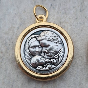 May include: Gold-toned pendant featuring a silver-toned image of a family. The image depicts a man, woman, and baby. The man and woman are gazing at the baby, while the baby looks up at the viewer.