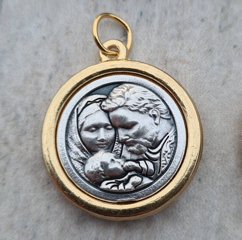 May include: Gold-toned pendant featuring a silver-toned image of a family. The image depicts a man, woman, and baby. The man and woman are gazing at the baby, while the baby looks up at the viewer.