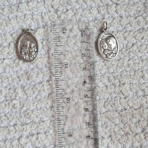 May include: Two silver oval-shaped pendants with a religious theme. The larger pendant features the words "Holy Family" and a depiction of Mary, Joseph, and baby Jesus. The smaller pendant depicts a group of three people. Both pendants are measured against a ruler.