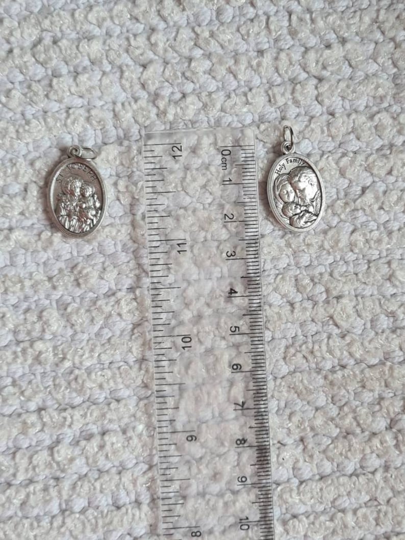 May include: Two silver oval-shaped pendants with a religious theme. The larger pendant features the words "Holy Family" and a depiction of Mary, Joseph, and baby Jesus. The smaller pendant depicts a group of three people. Both pendants are measured against a ruler.