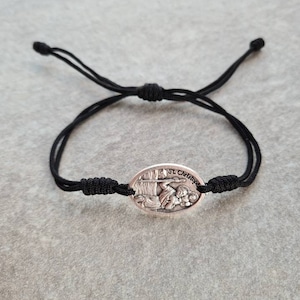 Könnte beinhalten: Ein schwarzes Kordelarmband mit einem silbernen ovalen Anhänger, der eine Darstellung des Heiligen Christophorus zeigt, der ein Kind trägt. Der Anhänger ist mit "St. Christopher" beschriftet.