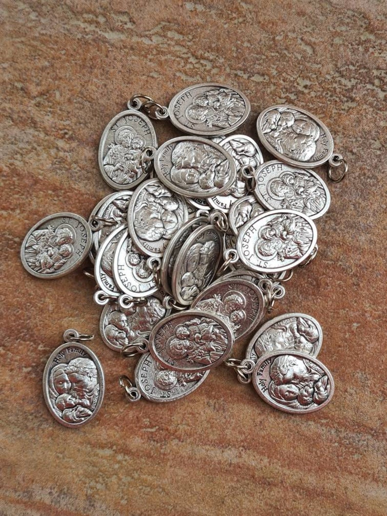 May include: A pile of silver-toned metal charms featuring an image of Saint Joseph, Mary, and baby Jesus. The charms are oval-shaped and have a loop at the top for attaching to a necklace or bracelet. The charms are all identical and feature the words "Holy Family" and "St. Joseph" on them.
