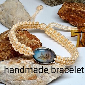 May include: A handmade bracelet with a cream-colored braided cord and a silver oval charm featuring a religious image. The bracelet has a wooden cross charm with the letters "INRI" on it.