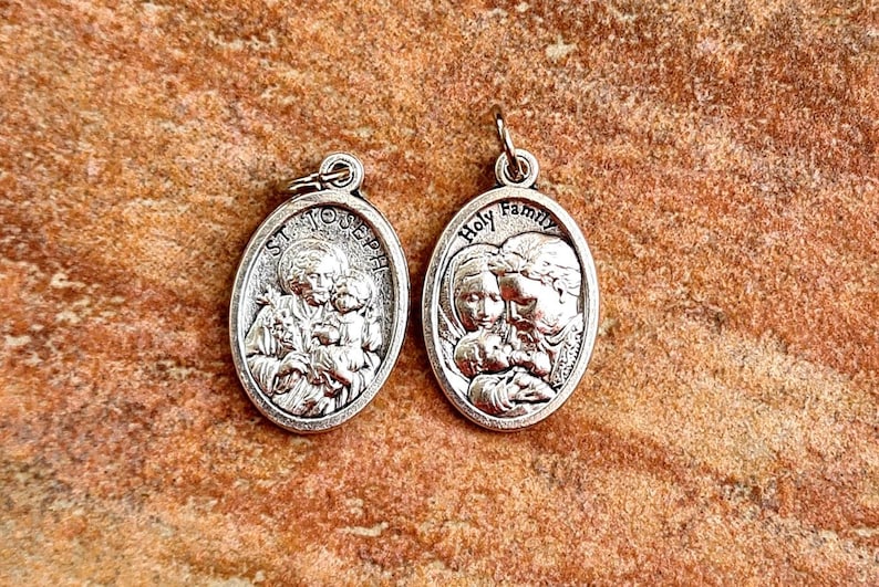 May include: Two silver oval-shaped medallions with religious imagery. The left medallion features a depiction of Saint Joseph holding the baby Jesus and Mary. The right medallion features a depiction of the Holy Family with Mary, Joseph, and the baby Jesus. Both medallions have a loop at the top for hanging.