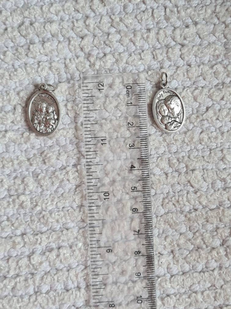 May include: Two silver oval-shaped pendants with a religious theme. The larger pendant features the words "Holy Family" and a depiction of Mary, Joseph, and baby Jesus. The smaller pendant depicts a group of three people. Both pendants are measured against a ruler.