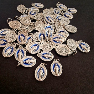 May include: A collection of silver-toned religious pendants with blue enamel detailing. The pendants feature images of a religious figure and text. Some pendants are oval, others are round, and all have a small loop for attaching to a chain.