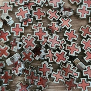 Puede incluir: Un montón grande de dijes de cruz de metal plateado con un diseño de esmalte rojo. Las cruces son todas del mismo tamaño y forma, y están dispuestas en un patrón aleatorio.