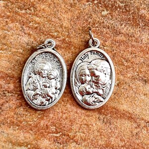 May include: Two silver oval-shaped medallions with religious imagery. The left medallion features a depiction of Saint Joseph holding the baby Jesus and Mary. The right medallion features a depiction of the Holy Family with Mary, Joseph, and the baby Jesus. Both medallions have a loop at the top for hanging.