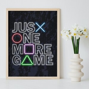 Może przedstawiać: Wydruk w ramie z czarnym tłem i białym tekstem, który brzmi "JUST ONE MORE GAME". Litery są obwiedzione neonowymi kolorami, a każda litera została zastąpiona innym symbolem przycisku kontrolera PlayStation. Wydruk wisi na białej ścianie obok wazonu z białymi kwiatami.