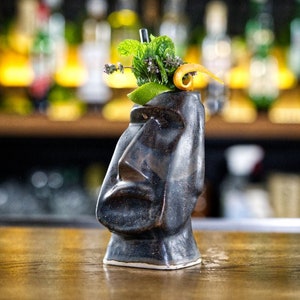 May include: A black ceramic tiki mug with a green leafy garnish and a curled orange peel. The mug is sitting on a wooden surface.