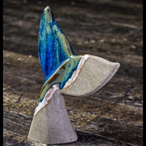Op de afbeelding: Een keramisch walvisstaartsculptuur met een blauwe, groene en witte glazuur. De staart is schuin omhoog gericht en de basis is lichtgrijs.