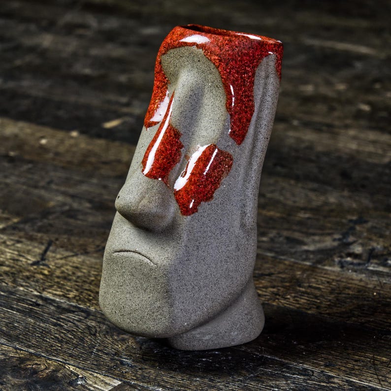 May include: A gray ceramic tiki mug with a red glaze. The mug is shaped like a human face with a large nose and a sad expression.