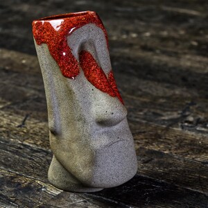 May include: A gray ceramic tiki mug with a red glaze on the top half. The mug is shaped like a face with a large nose and a small mouth.