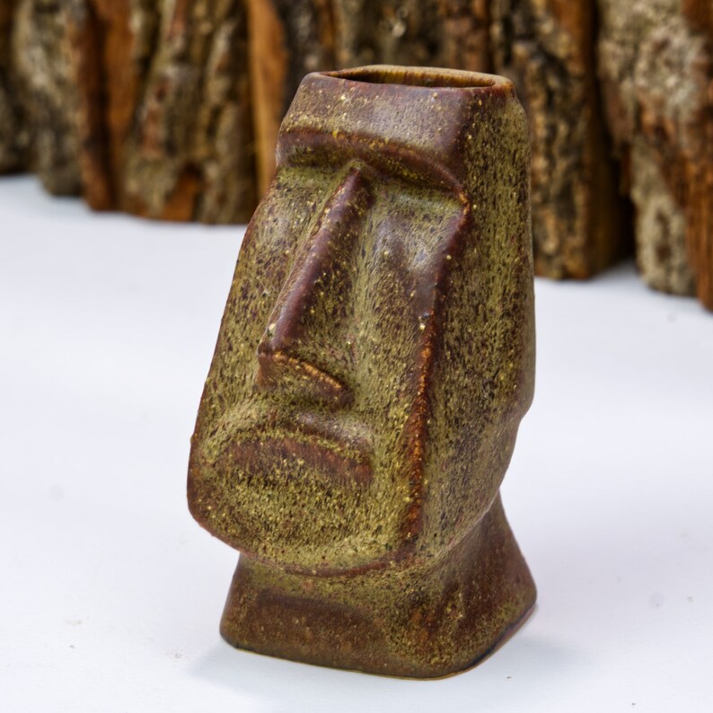 May include: A brown and green ceramic mug shaped like a traditional Easter Island moai statue. The mug has a textured surface and a detailed face.