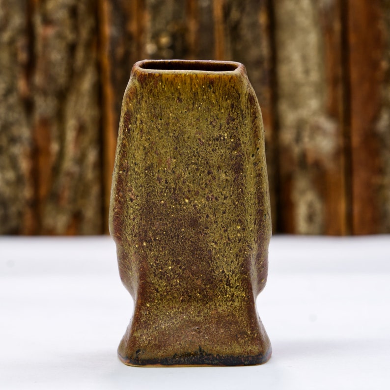 May include: A small, dark green and brown ceramic vase with a textured surface. The vase has a rectangular shape with rounded edges.