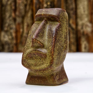 May include: A brown and green ceramic sculpture of a Moai head. The sculpture has a textured surface and a simple, geometric design.