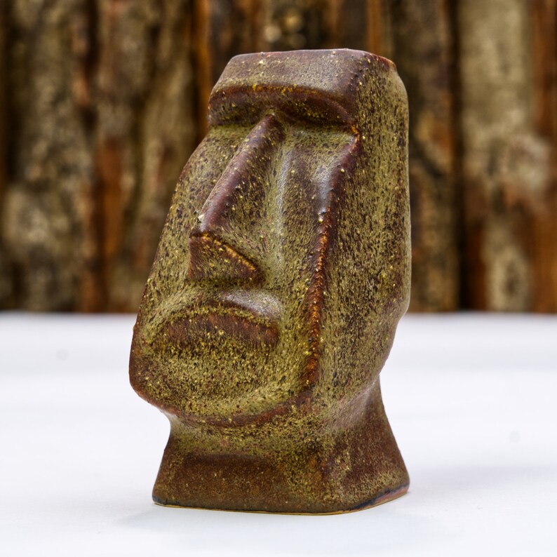 May include: A brown and green ceramic sculpture of a Moai head. The sculpture has a textured surface and a simple, geometric design.