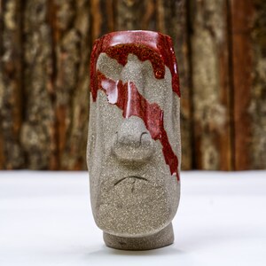 May include: A gray ceramic mug shaped like a face with red glaze dripping down the side. The mug has a large nose and a small mouth.