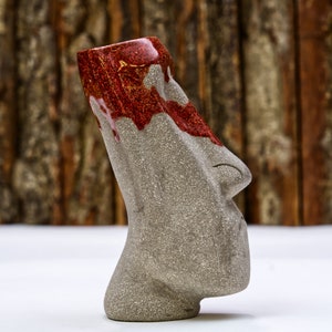 May include: A gray ceramic tiki mug with a red and gold glaze. The mug has a human-like face with a nose and mouth.