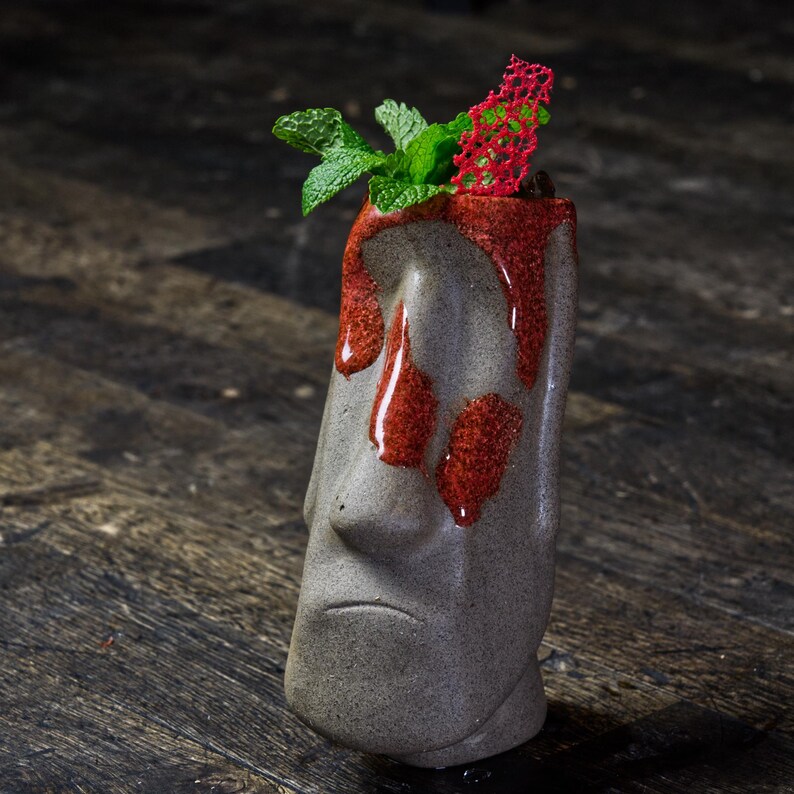 May include: A gray ceramic tiki mug with a red glaze, topped with a sprig of green mint and a red decorative element.