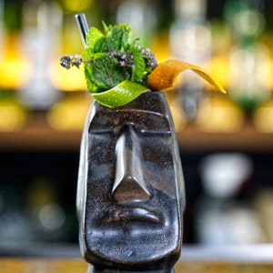 May include: A dark gray ceramic tiki mug with a face sculpted on the front. The mug is topped with a sprig of green mint and a slice of orange peel.