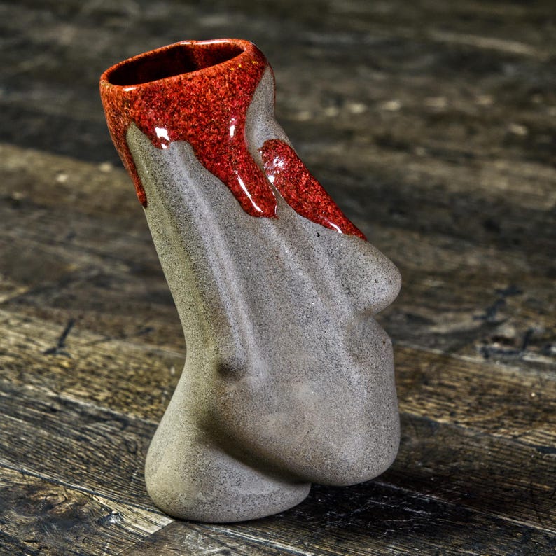 May include: A gray ceramic mug with a red glaze dripping down the side. The mug is shaped like a face with a prominent nose.