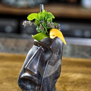 May include: A black ceramic tiki mug with a face, filled with a green and purple drink, garnished with a lime wedge and a sprig of mint.