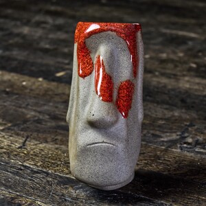 May include: A gray ceramic tiki mug with a red glaze dripping down the front. The mug has a carved face with a nose and a mouth.