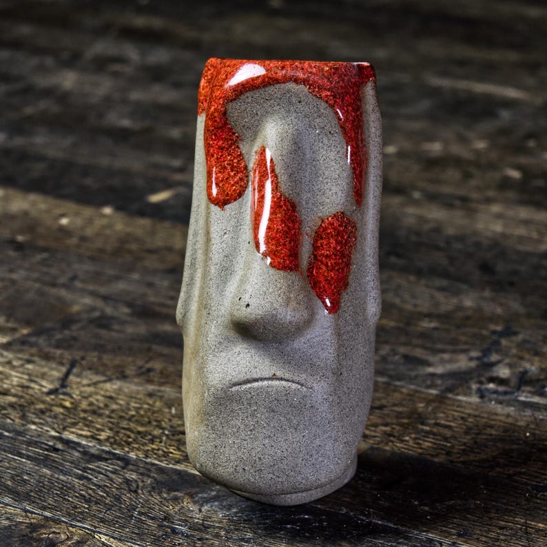 May include: A gray ceramic tiki mug with a red glaze dripping down the front. The mug has a carved face with a nose and a mouth.