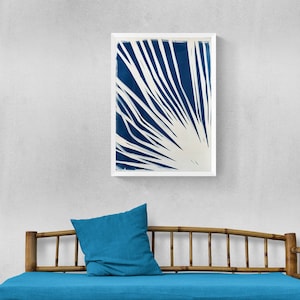 May include: A blue and white abstract print in a white frame hangs above a bamboo daybed with a blue cushion.
