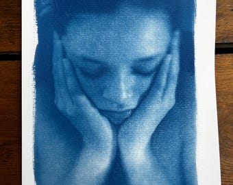May include: A cyanotype photograph of a person with their head resting on their hands. The image is in focus and the person's face is visible.