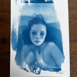 Pode incluir: Uma fotografia de cianotipia de uma pessoa numa banheira. O rosto da pessoa é visível e está a olhar diretamente para a câmara. A imagem tem um aspeto desbotado e vintage.