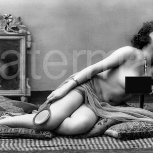 Op de afbeelding: Een vintage zwart-witfoto van een vrouw die op kussens ligt en een handspiegel vasthoudt. Ze is gehuld in doorzichtige stof en draagt een parelketting. Een decoratieve spiegel en stoel zijn op de achtergrond.