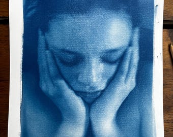 May include: A cyanotype photograph of a person with their chin resting on their hands. The person's face is obscured by their hands, but their eyes are closed.