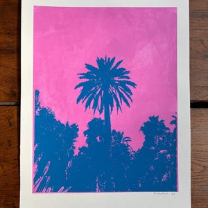 Peut inclure: Une estampe représentant une silhouette de palmier bleu sur fond rose. L'œuvre d'art comprend une représentation détaillée des frondes du palmier et du feuillage environnant. La signature de l'artiste et le numéro 25 sont visibles en bas à droite.